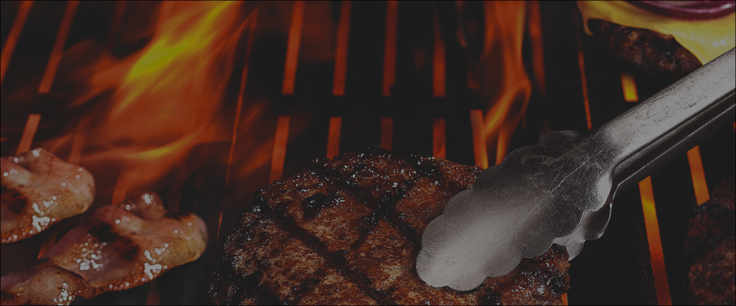 Silver tongs turning a piece of chicken on flaming grill with tomatoes and corn on the cob.