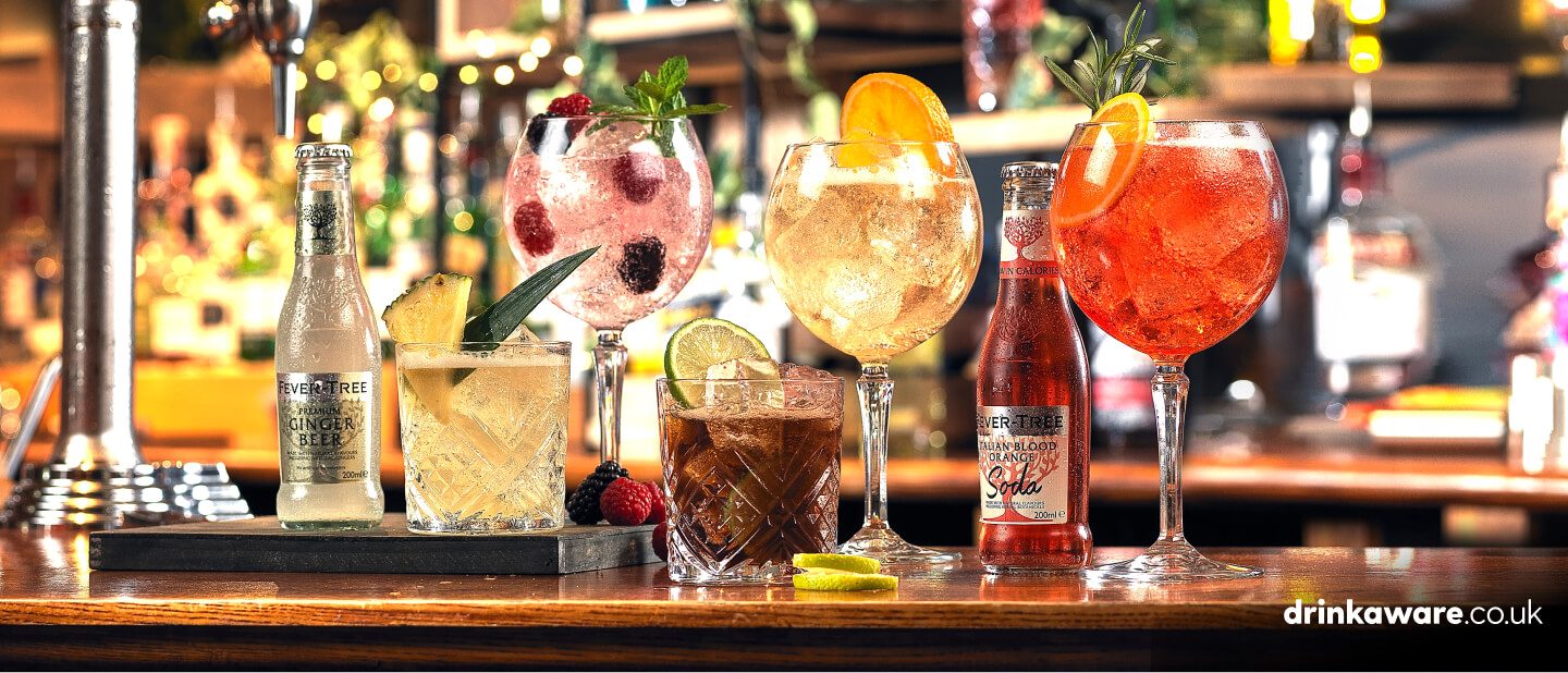 A selection of Beefeater drinks on a bar with raspberries, lime and pineapple garnishes and bottles of ginger beer and blood orange soda.